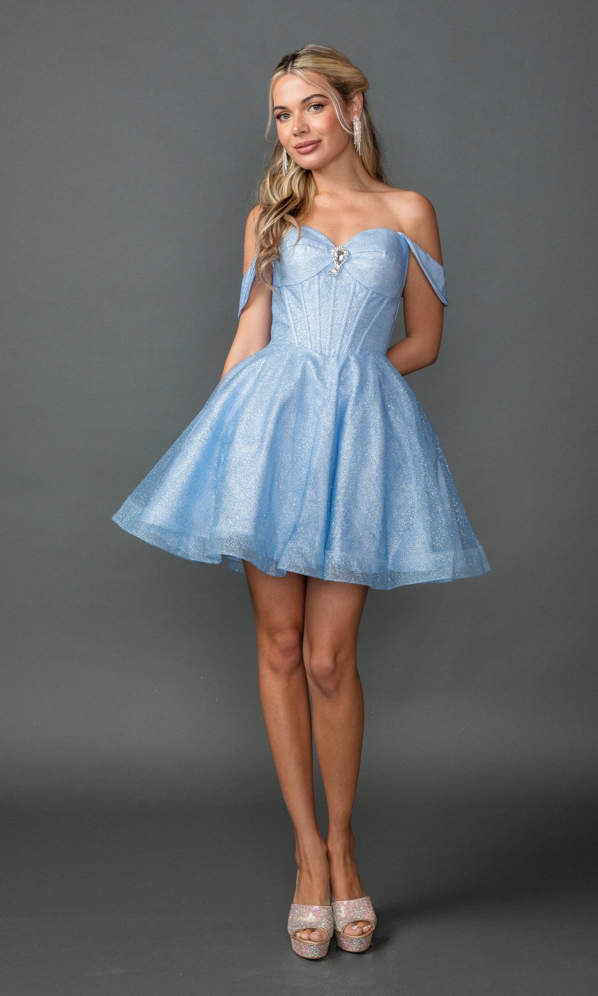 A woman poses against a gray background wearing the Dancing Queen 3425 short prom dress in light blue, featuring an off-the-shoulder neckline, flared glitter tulle skirt, and paired with silver high heels.